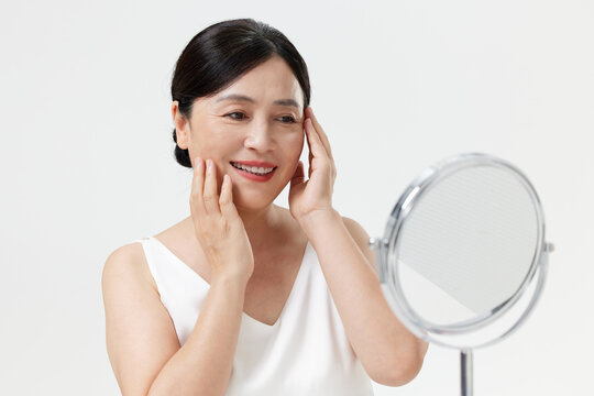 Happy mature Asian woman touching face and looking in mirror for skincare