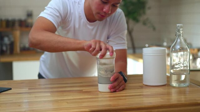 Asian man preparing protein shake at home kitchen counter with shaker bottle, protein powder container, and glass water bottle