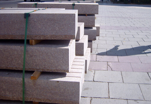 Stack of heavy granite curb stones secured with green straps on wooden spacers, placed on a paved sidewalk during urban road construction in bright sunlight.