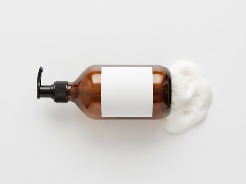 Top down flat lay view of an amber glass pump bottle with blank white paper label lying horizontally with white soap foam at the base isolated on white background. Skincare packaging mockup.