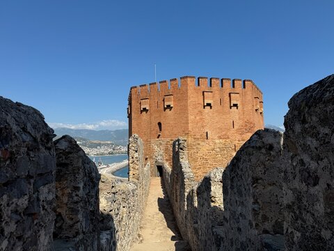 Scenic view of the historic Red Tower (Kızılkule) from inside the ancient fortress walls in Alanya, Turkey, featuring a blue sky, stone walkway, and coastal harbor background.
