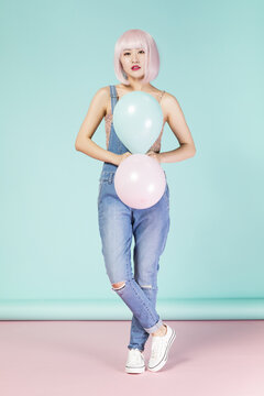 Young Woman in Pink Wig and Denim Overalls Holding Pastel Balloons in Studio