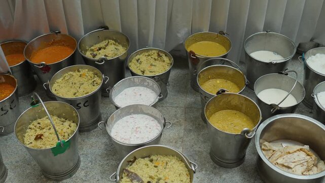 Metal buckets packed with South Indian wedding dishes including curries, rice, raita and breads arranged on the floor.