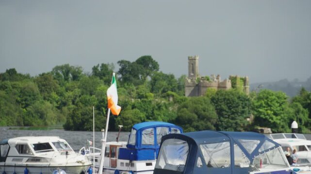 An Irish tricolour is displayed above moored pleasure boats on Lough Key, with McDermott's Castle on wooded Castle Island in the background.