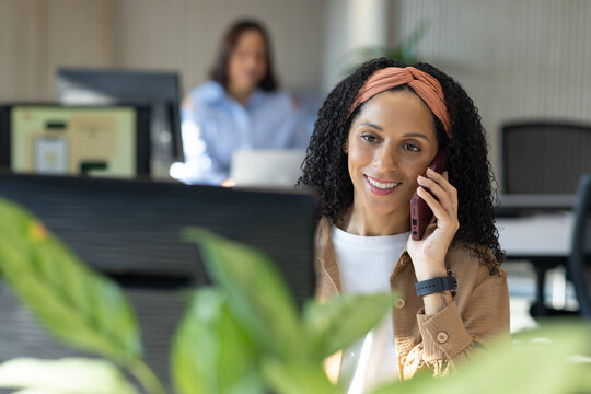 Female coworkers talking while woman using smartphone at desk facing monitor wearing brown shirt