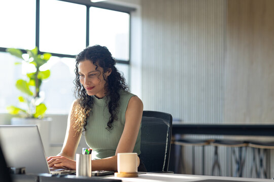 Woman typing on laptop at office desk, wearing sleeveless green top, mug, pen cup, plant