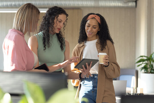 Diverse female coworkers standing together around tablet in modern office, pointing at screen