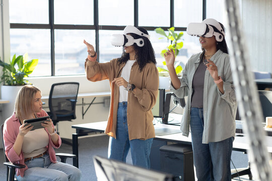 Three female coworkers using white VR headsets, gesturing while peer holding tablet in office