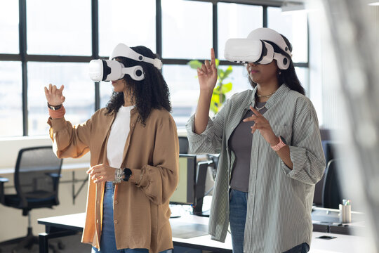 Diverse female coworkers interacting with virtual controls in open office wearing white VR headsets