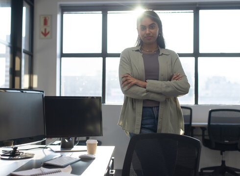 Indian woman wearing button-up and jeans, standing in open office near dual monitors, coffee cup