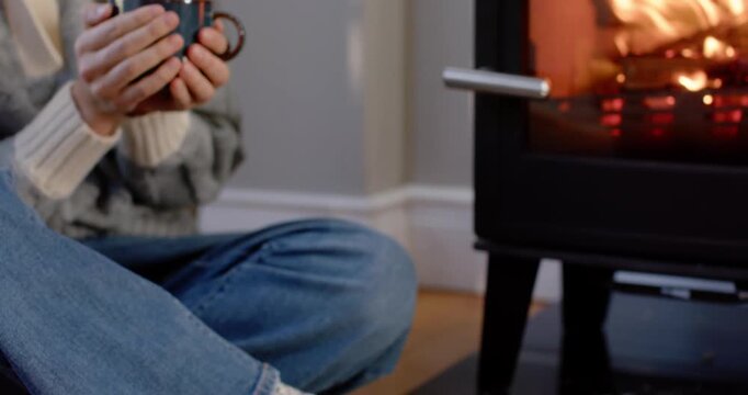 Woman sitting on wood floor by wrapped gifts, holding ceramic mug, seeking warmth from stove