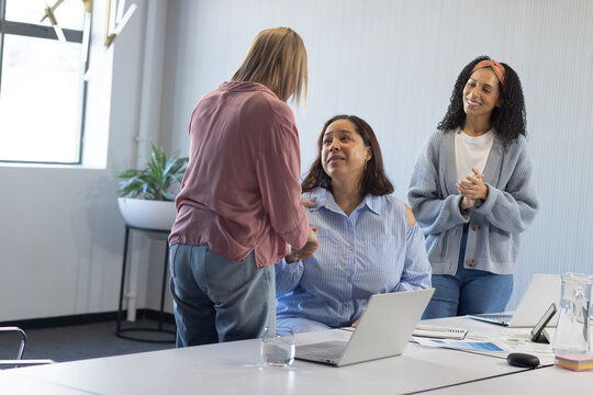 Diverse female colleagues shaking hands and clapping at conference table with laptops and charts