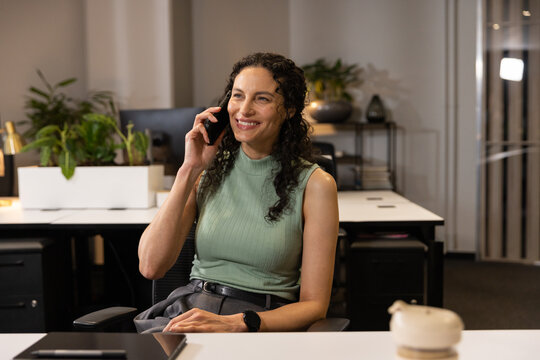 African American female sitting at office desk smiling while holding smartphone wearing green top