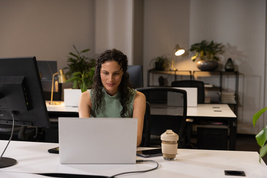 Woman wearing sleeveless top working on silver laptop in office with monitor and smartphone