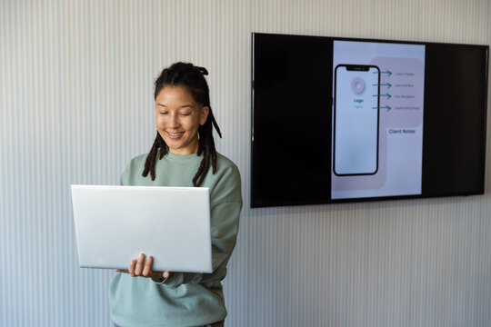 Nonbinary designer holding silver laptop reviewing phone UI on screen in office mint top