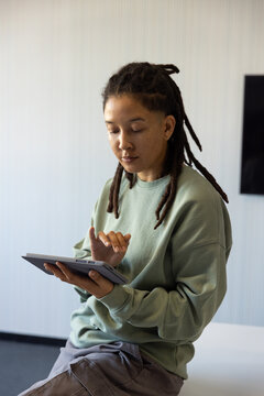 African nonbinary person sitting on table edge wearing sage sweater, tapping tablet folio in office