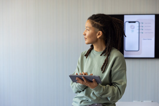 Non-binary adult in green top seated at office holding tablet, showing app on monitor, copy space