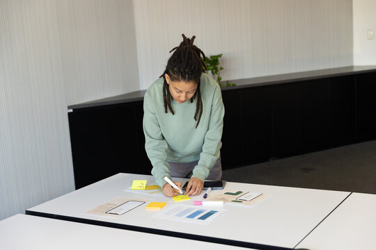 Nonbinary African American leaning over modular table in office, writing on yellow sticky note
