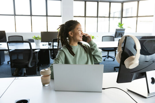 African American nonbinary adult in green sweatshirt speaking on phone at office desk with laptop