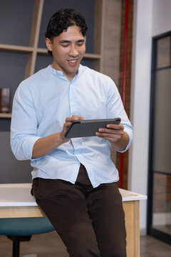 Asian man sitting on light wood desk holding tablet smiling in office wearing light blue shirt