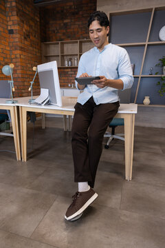 Man sitting on wooden desk in modern office wearing light blue shirt, holding tablet