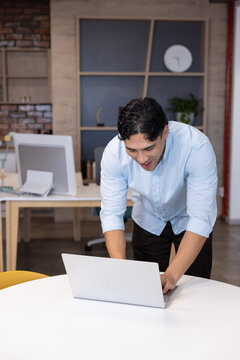 Asian man leaning over silver laptop at round table in modern office, wearing light-blue shirt