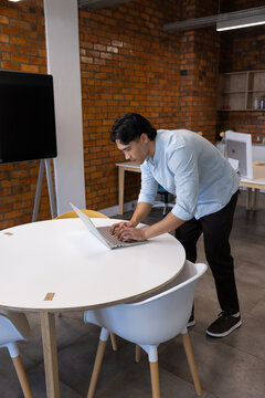 Asian man wearing light blue shirt leaning over round white table typing on laptop at office