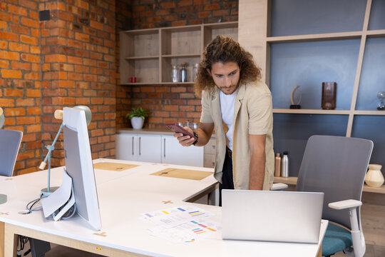 Adult man leaning over shared desk in modern workspace, checking silver laptop and smartphone