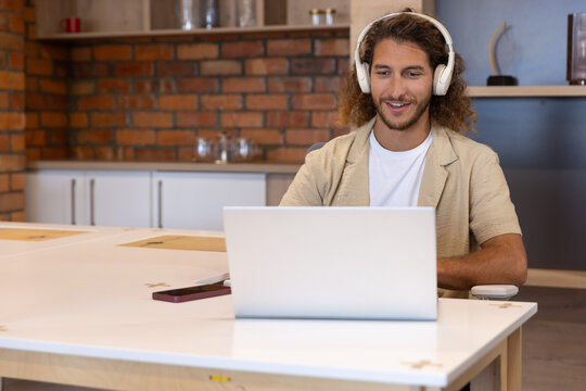 Man sitting at white table in kitchen wearing white headphones using laptop and smartphone