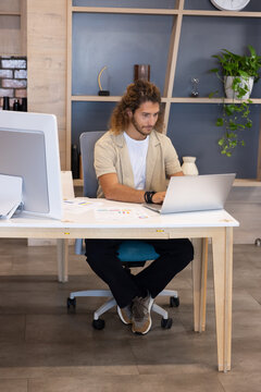 Adult male sitting at white office desk working on silver laptop and scanning printed charts
