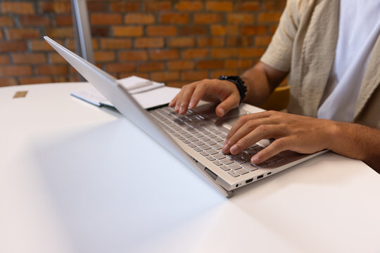 Man in his twenties typing on silver laptop at desk with spiral notebook, wearing beige overshirt