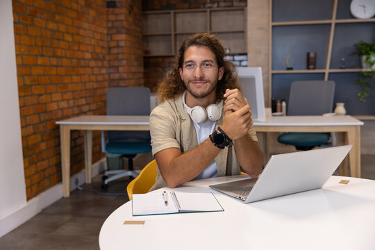 Man sitting at round white table in co-working studio using silver laptop, notebook and headphones