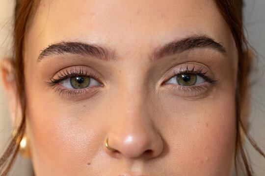 Female facing camera in studio, showing hair pulled back, gold nose hoop, mascara, foundation