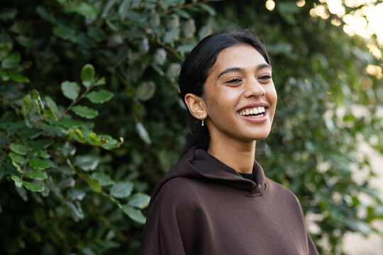 Indian woman smiling, standing and turning at hedge, wearing dark brown hoodie with gold earrings