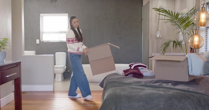 Asian woman walking into bedroom carrying box, unpacking folded clothing on bed by tub, copy space