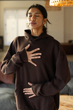 Female standing with eyes closed practicing slow breathing in living room wearing dark brown hoodie