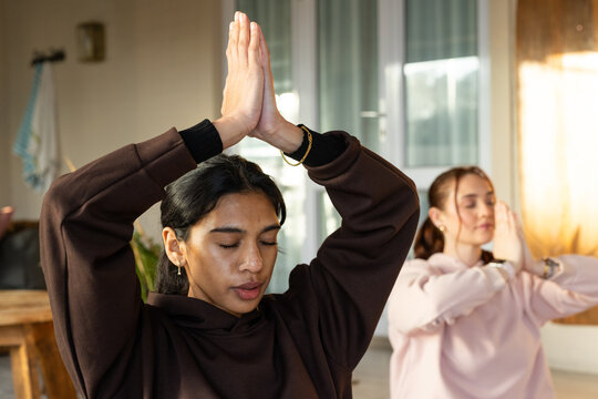 Diverse female friends practicing meditation at home in hoodies near glass doors and wooden table