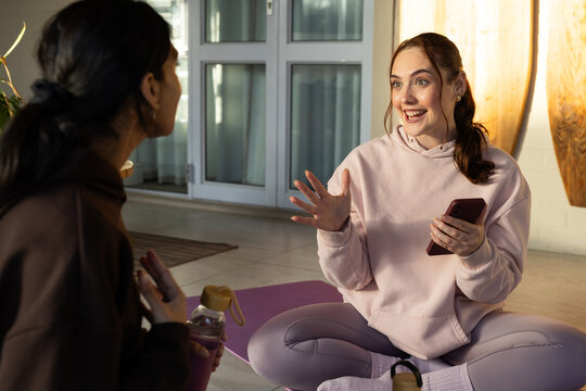 Diverse female friends in hoodies sitting on yoga mats in bright room, chatting and holding phone