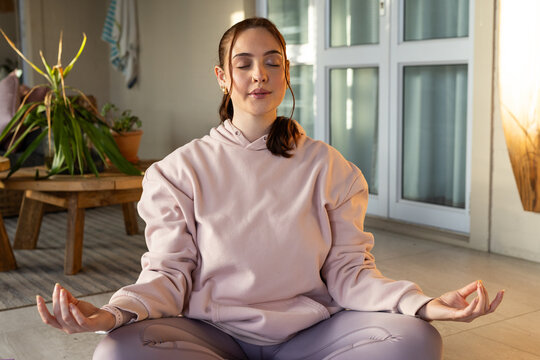 Female sitting cross-legged meditating at home wearing pink hoodie and leggings with potted plants