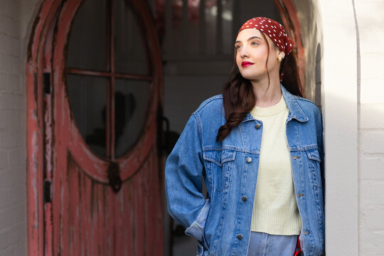 Woman standing in arched entryway beside weathered red round door, wearing denim jacket, headscarf