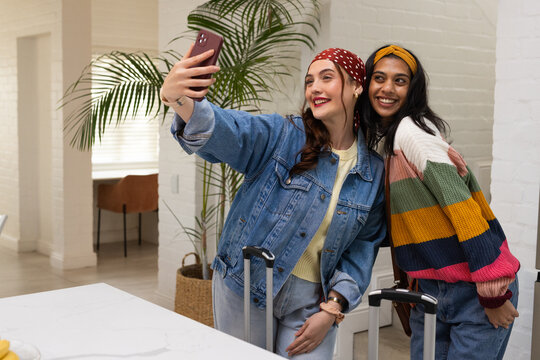 Diverse female friends in denim and stripes posing, taking selfie with phone and suitcases in entry