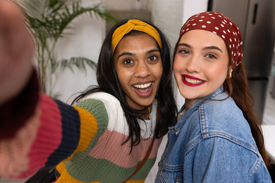 Diverse female friends posing together, taking selfie in kitchen, holding smartphone and headscarf