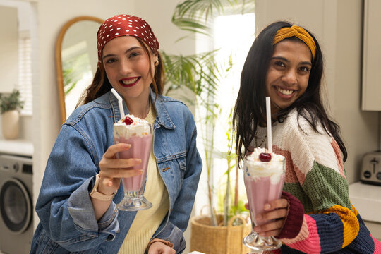Diverse female friends standing at kitchen counter holding milkshakes with cream and cherry, washer