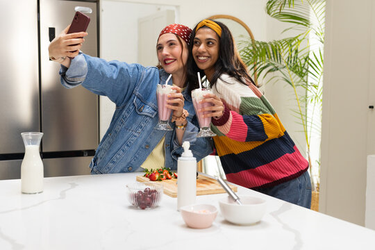 Diverse female friends taking selfie in kitchen wearing denim jacket holding milkshakes, smartphone