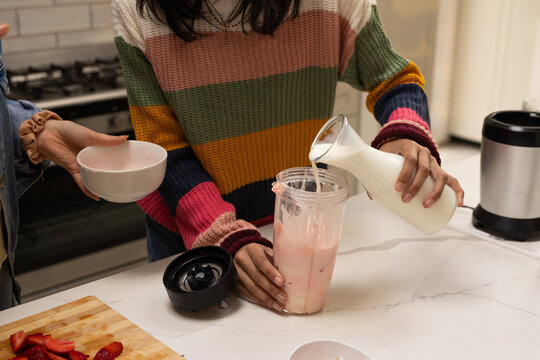 Diverse women wearing striped pouring from pitcher into blender cup making shake on kitchen counter
