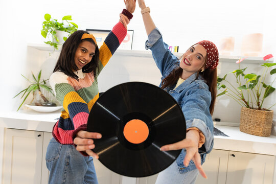 Diverse female friends, one wearing denim jacket, high-fiving and holding orange vinyl in kitchen