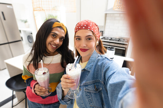 Two adult women in stripes and denim holding milkshakes at kitchen island, taking selfie with phone