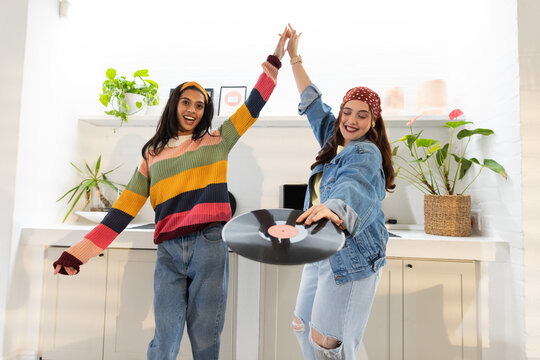 Diverse women dancing and holding vinyl record in home kitchen, wearing yellow headband