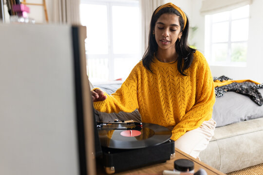 Indian woman in mustard knit sitting on sofa operating black turntable with vinyl in bright bedroom