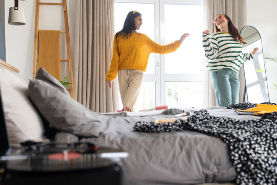 Diverse female friends dancing and moving in bedroom with bed and record player near glass door